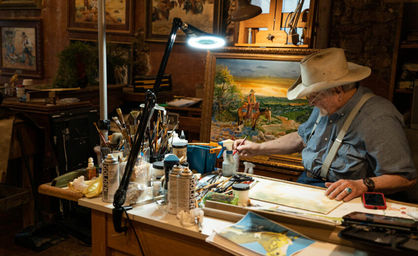 Western artist Jack Moss sits at his work desk surrounded by canvases he has coated in iconic Western imagery. Moss is a self-taught Western artist who set up shop at Gridiron Gallery in downtown Llano. Staff photo by Dakota Morrissiey