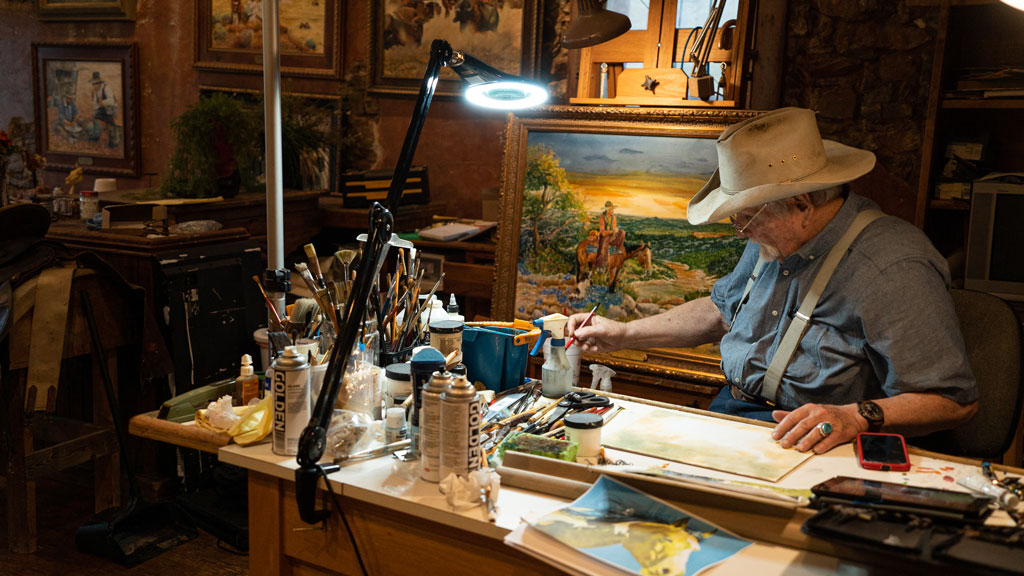 Western artist Jack Moss sits at his work desk surrounded by canvases he has coated in iconic Western imagery. Moss is a self-taught Western artist who set up shop at Gridiron Gallery in downtown Llano. Staff photo by Dakota Morrissiey